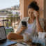 Eine Frau sitzt sichtbar angeschlagen auf dem Balkon ihrer Ferienunterkunft und blickt besorgt auf ihr Smartphone. Neben ihr stehen ein Laptop, Reisegepäck und Taschentücher, was den Eindruck vermittelt, dass sie sich während ihres Urlaubs mit einer Krankschreibung beschäftigt.